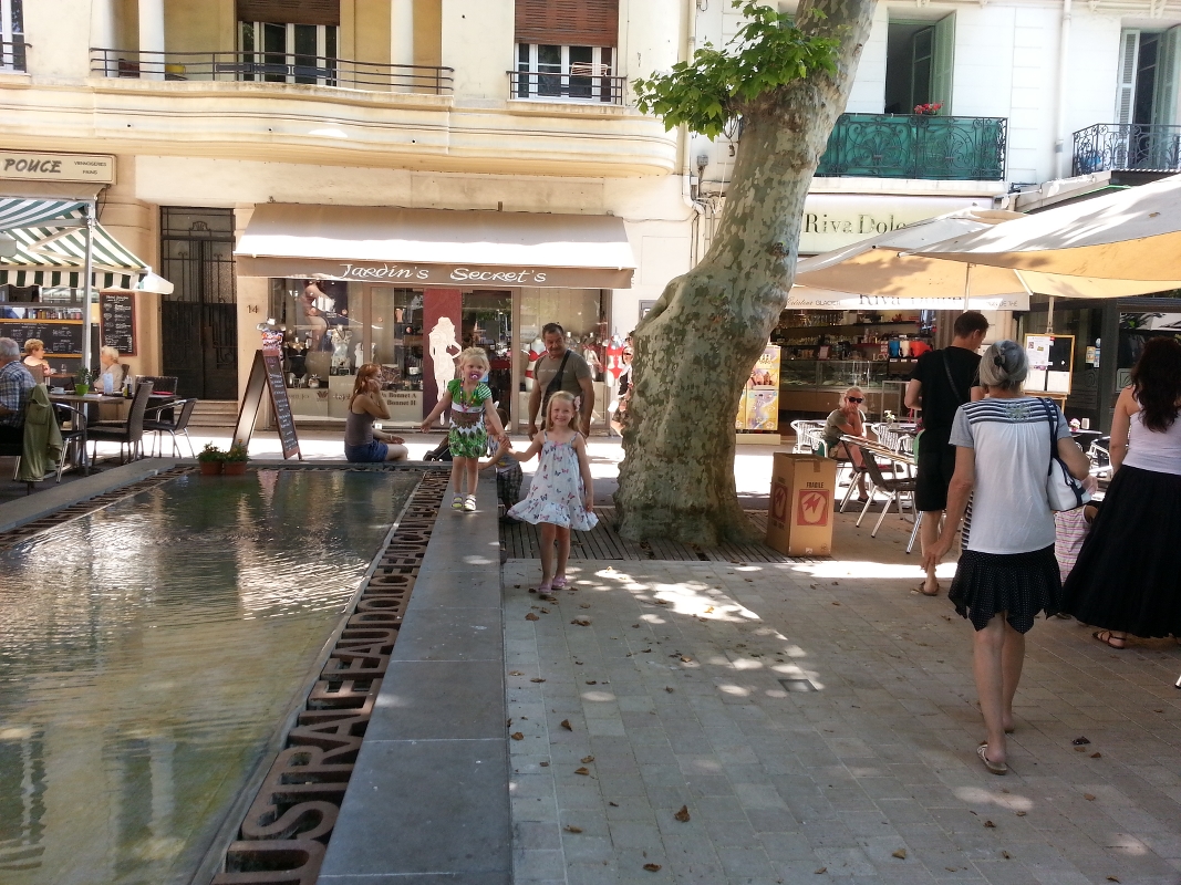 Folk spaserer og slapper av ved en grunn fontene på et solfylt utendørs torg med kafeer og butikker i bakgrunnen.