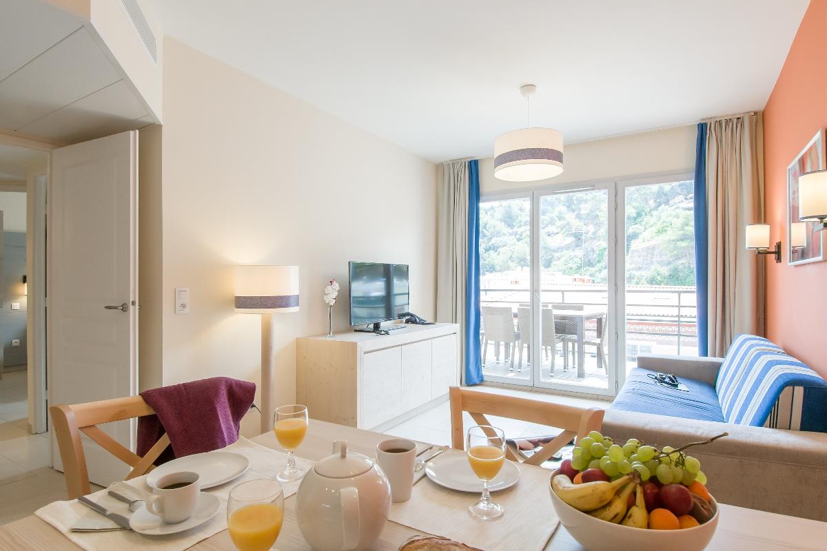 Lys stue og spisestue med dekket frokostbord, sofa, TV og balkongdører med fjellutsikt.