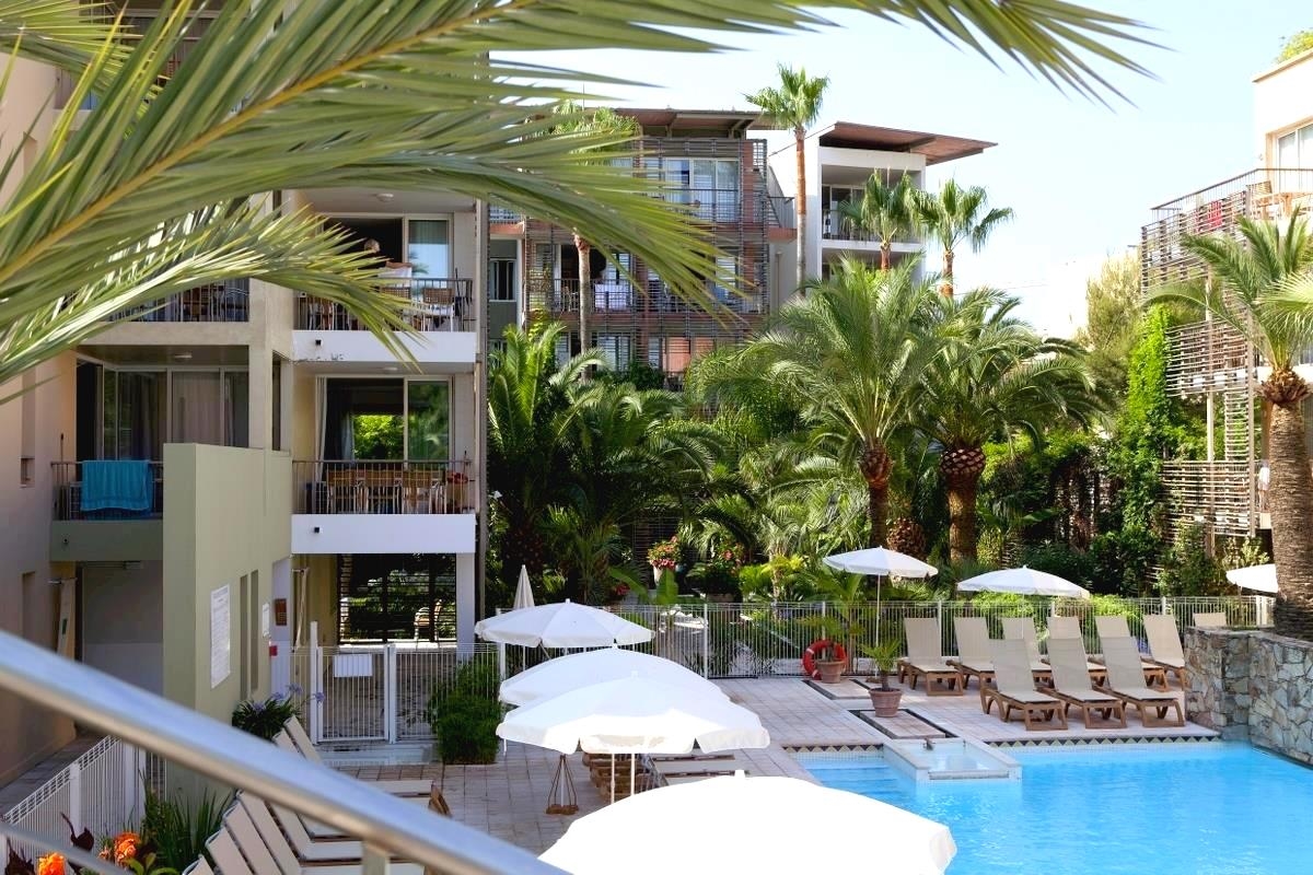 Utendørsbasseng med solsenger, hvite parasoller og frodige palmer ved siden av en moderne hotellbygning.