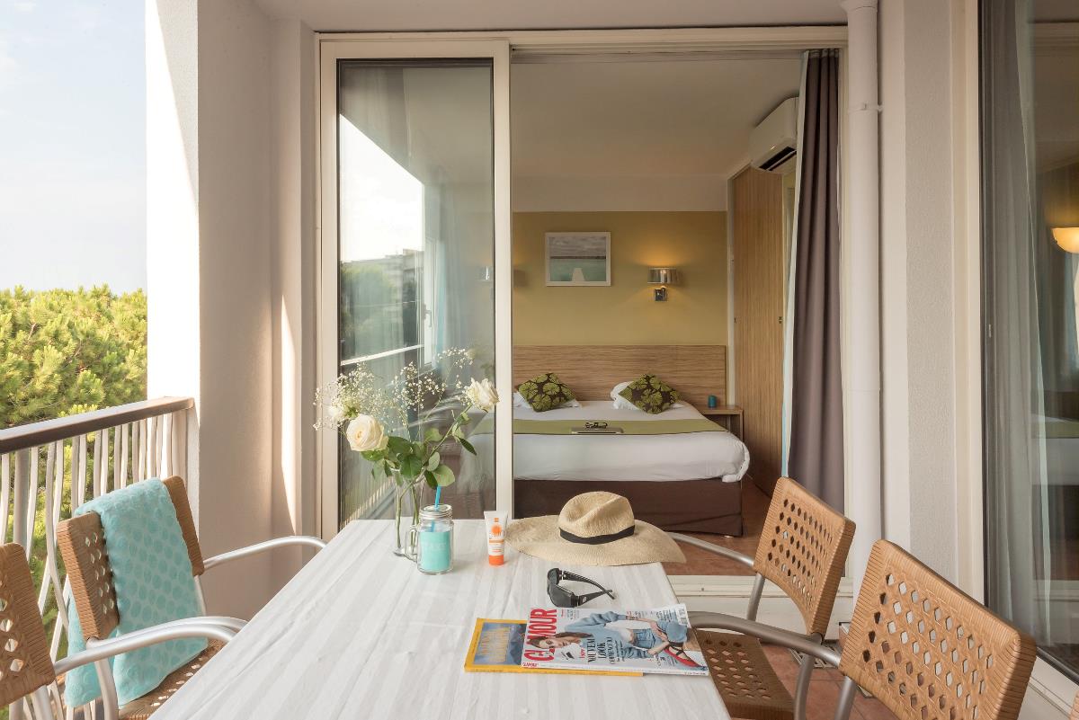 Et koselig hotellrom med balkong, bord, stoler, magasiner, blomster, hatt og solbriller på en solrik dag.
