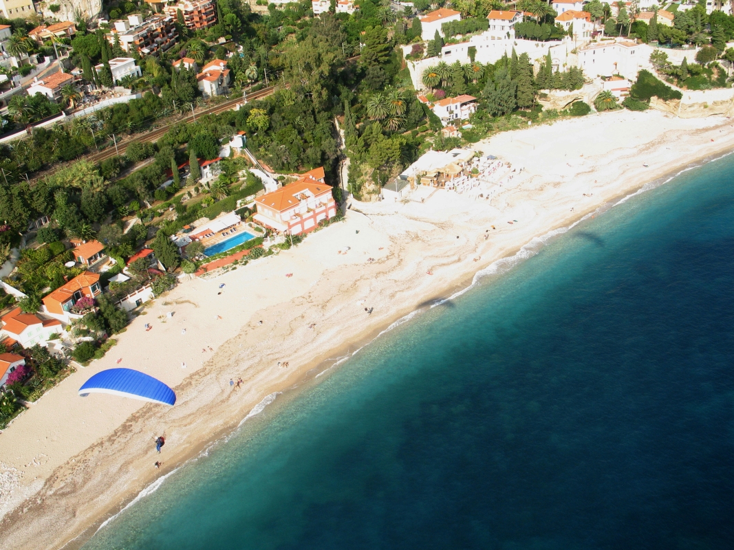 En person paraglider over en sandstrand nær klart, blått vann, med hus og grøntområder langs kysten.