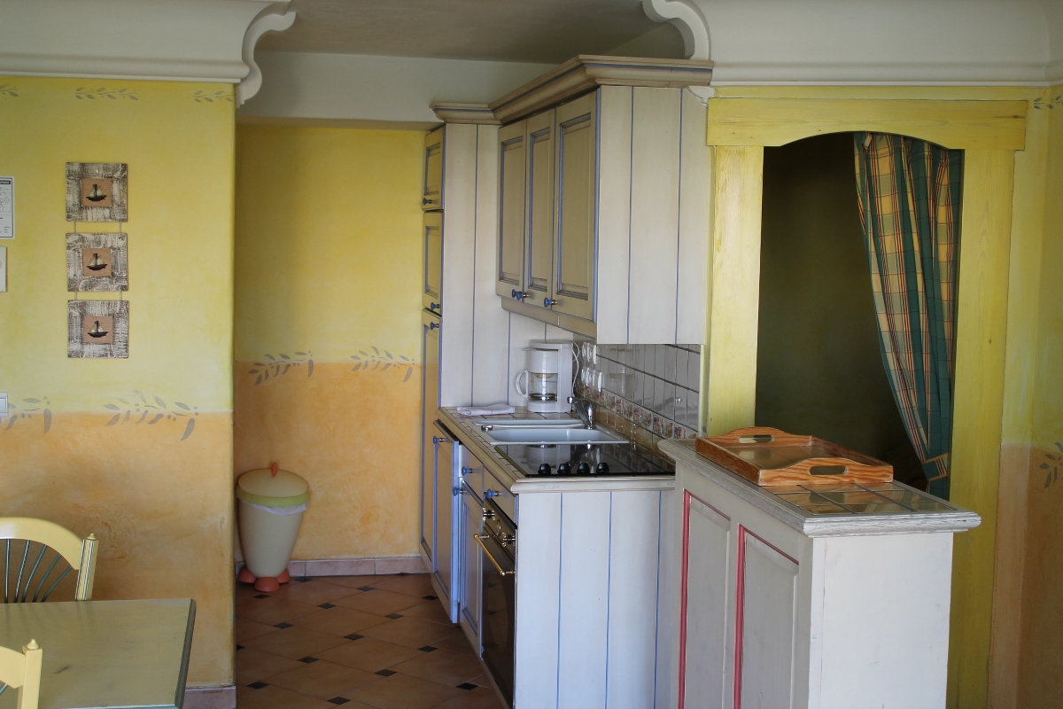 Koselig kjøkken med lysegule vegger, flislagt backsplash, komfyr, skjærebrett og rutete gardin over en buet døråpning.