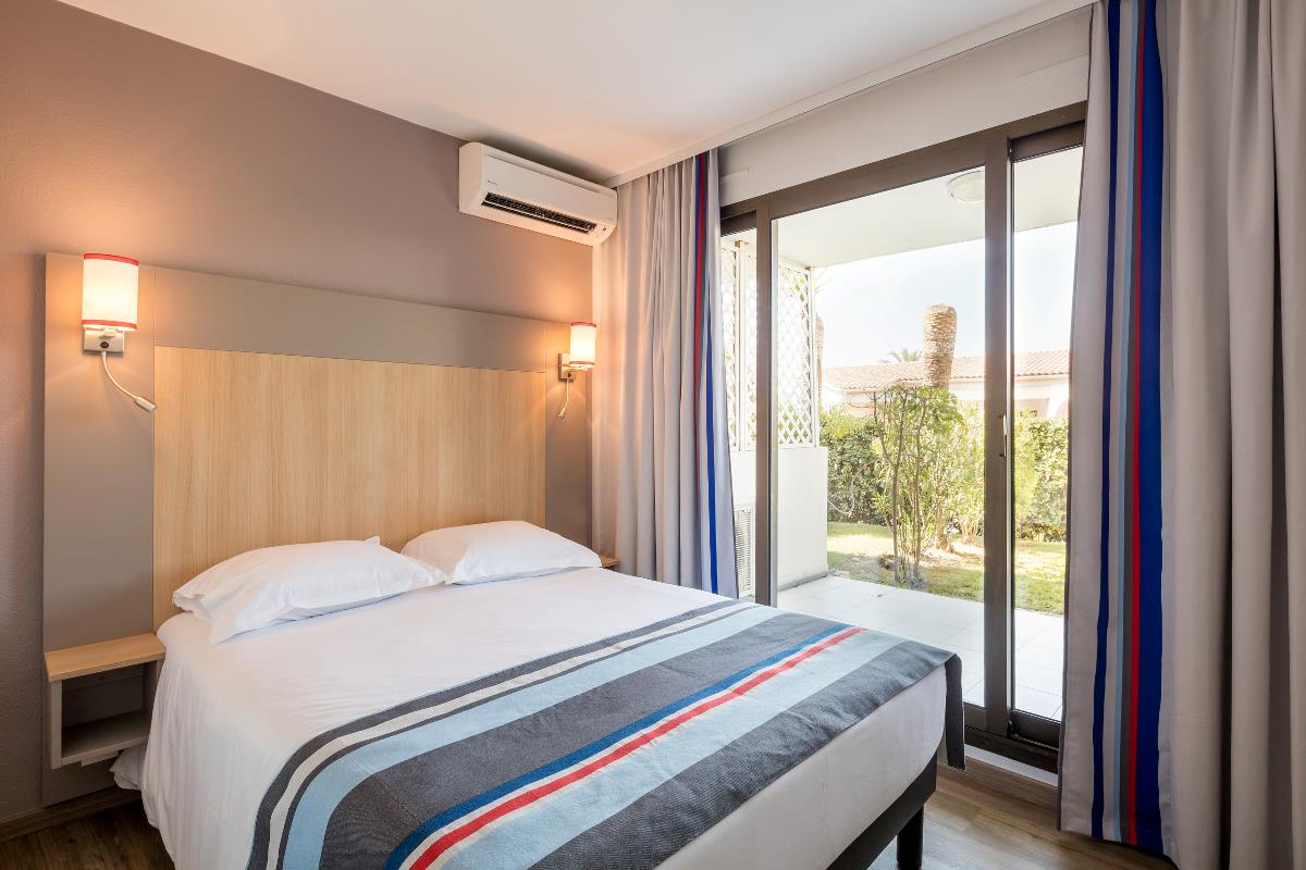 Et moderne hotellrom med dobbeltseng, stripete sengetøy og skyvedører i glass som fører ut til en uteplass i hagen.