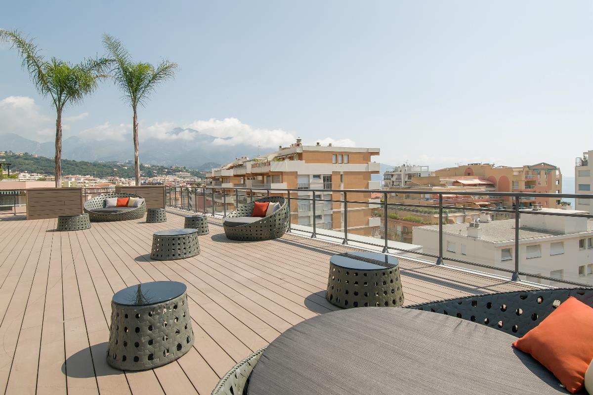 Moderne takterrasse med kurvmøbler, palmer og utsikt over byen under en lys, lett overskyet himmel.