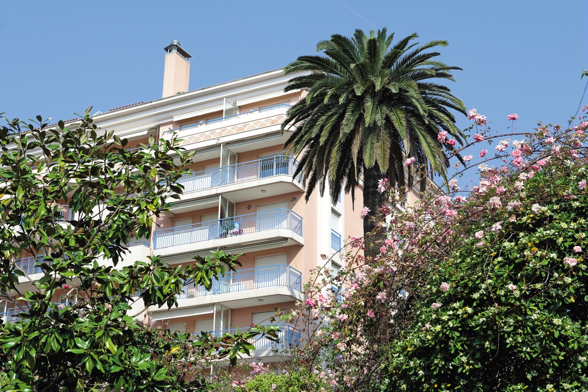 Leilighetsbygning med balkonger, palmer og blomstrende planter foran, under en klar blå himmel.