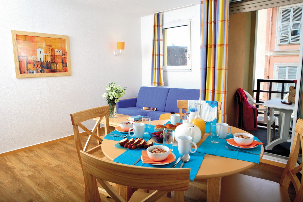 Et spisebord dekket til frokost i en lys stue med en blå sofa og fargerike gardiner.