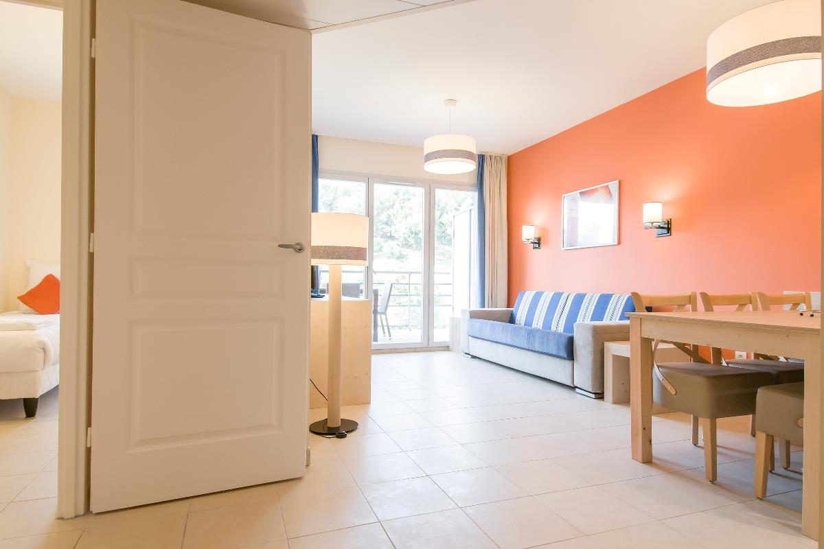 Lys stue og spisestue med oransje vegg, blå sofa, bord og åpen dør til et soverom.