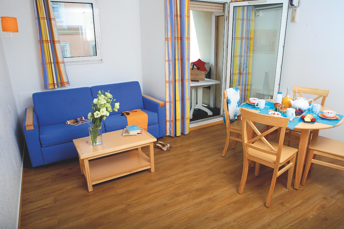 Lys stue og spiseplass med blå sofa, trebord, stoler, blomster og frokost dekket på bordet.