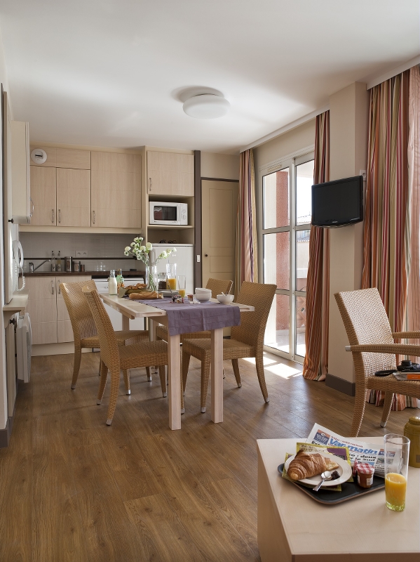 Moderne kjøkken og spiseplass med tregulv, kurvstoler, frokostbord og sollys som strømmer inn gjennom vinduene.