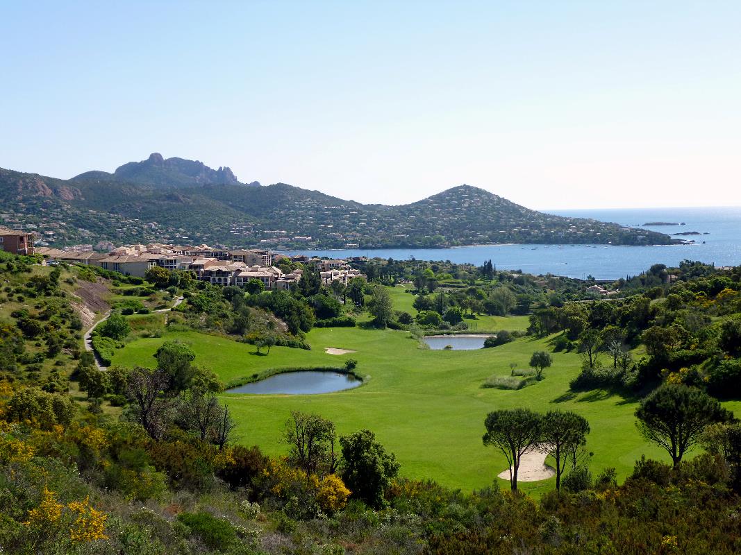Grønn golfbane med dammer, omgitt av åser, bygninger og et fjernt hav under en klar himmel.