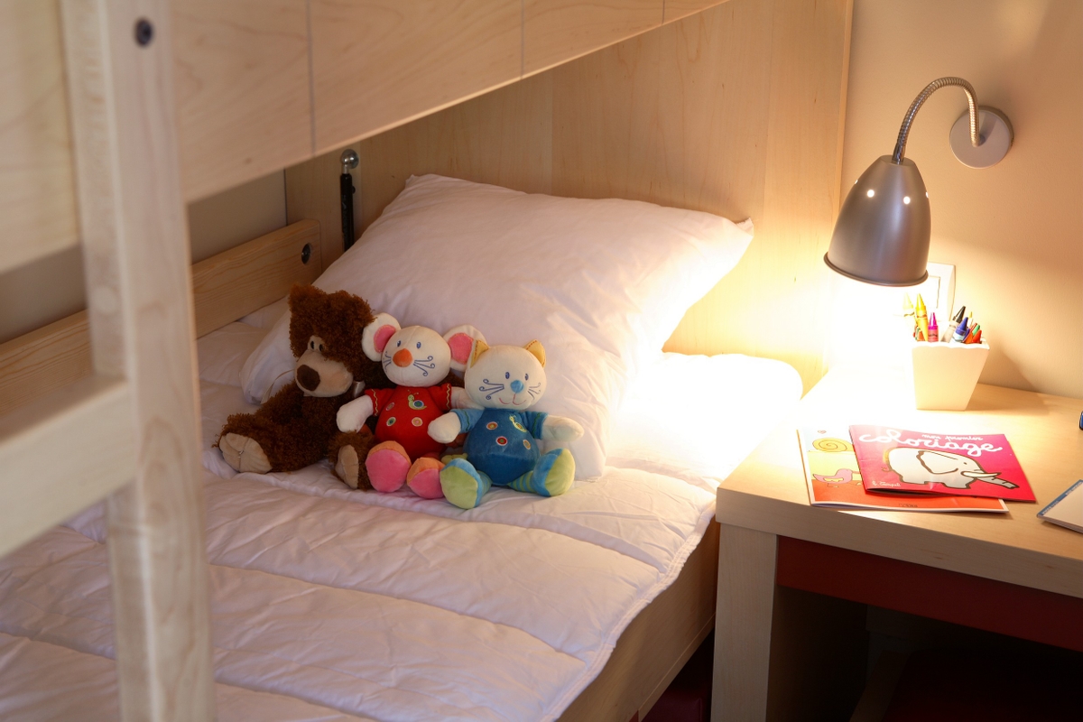 Kosebamse, mus og katt på en pent oppredd seng ved siden av et skrivebord med lampe, bøker og fargeblyanter.