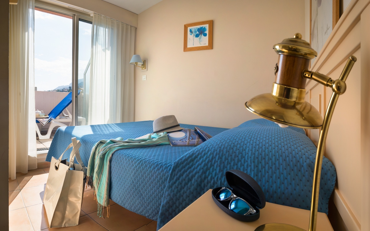 Solfylt hotellrom med blått sengeteppe, hatt, bok og skjerf på sengen, og solbriller på et bord ved en lampe.