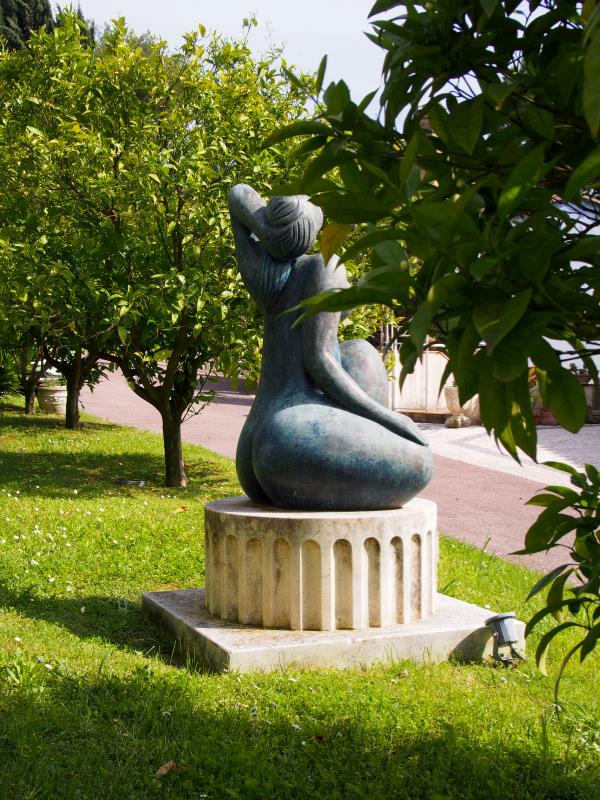 Svinget abstrakt skulptur av en sittende figur på en sokkel, omgitt av frodige grøntområder og trær på en solrik dag.