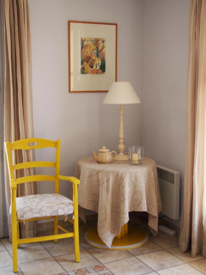 En gul stol og et rundt bord med lampe, tekanne og glass står i et koselig, solfylt hjørne med beige gardiner.