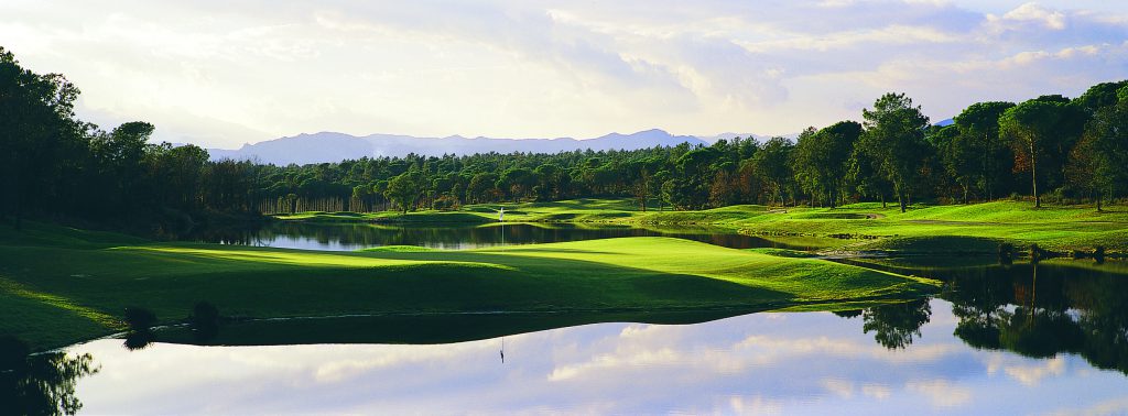 Vakker utsikt over en golfbane med frodige grønne fairways, som reflekterer vannhinder, trær og fjerne fjell under en overskyet himmel.