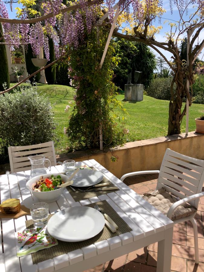 Et solfylt utendørsbord dekket for to med salat, vann og blomster over hodet, med utsikt over en frodig, grønn hage.