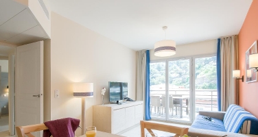 Lys stue med sofa, TV, spisebord og store vinduer ut mot en balkong med naturskjønn utsikt.