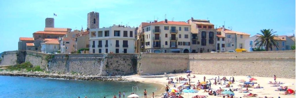Folk slapper av på en sandstrand nedenfor gamle steinbygninger og en mur i en kystby ved Middelhavet under en klar himmel.
