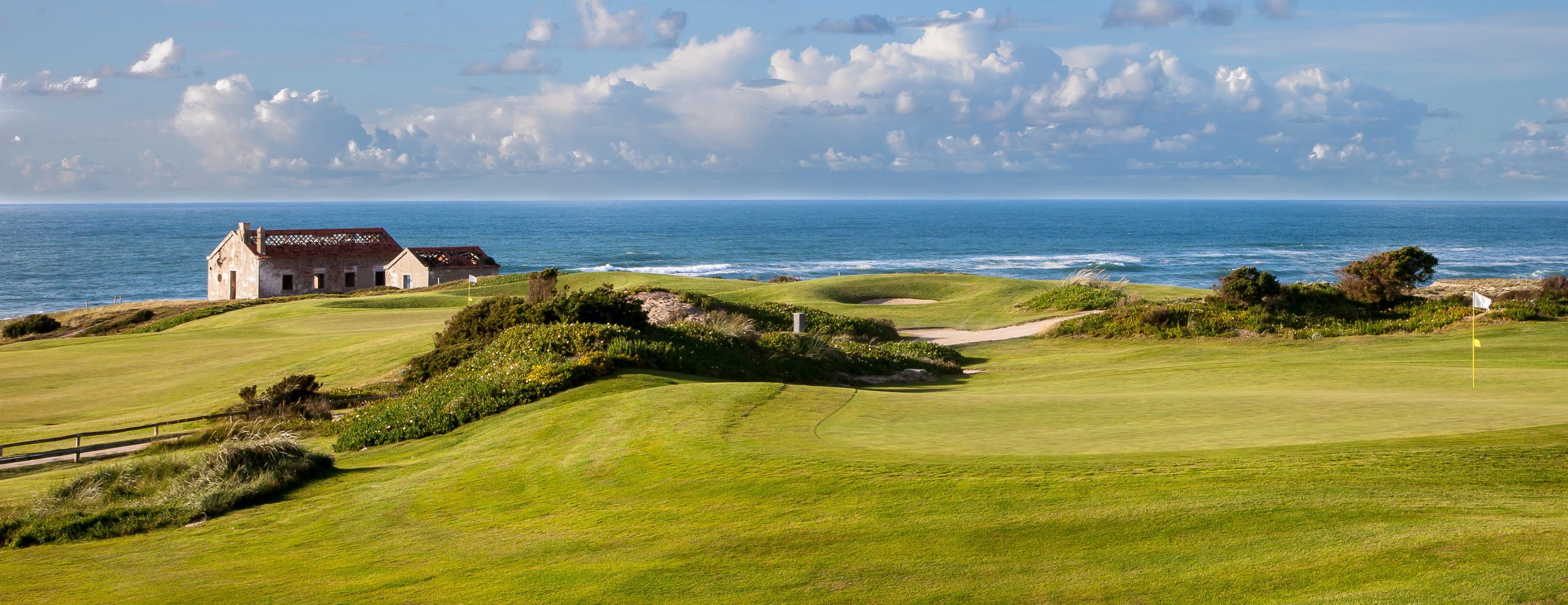 Kystgolfbane med bølgende greener, en gammel steinbygning og havbølger under en delvis overskyet himmel.