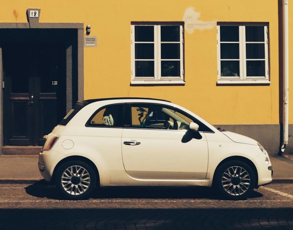 En liten hvit bil parkert foran en gul bygning med flere vinduer og en sentral døråpning.