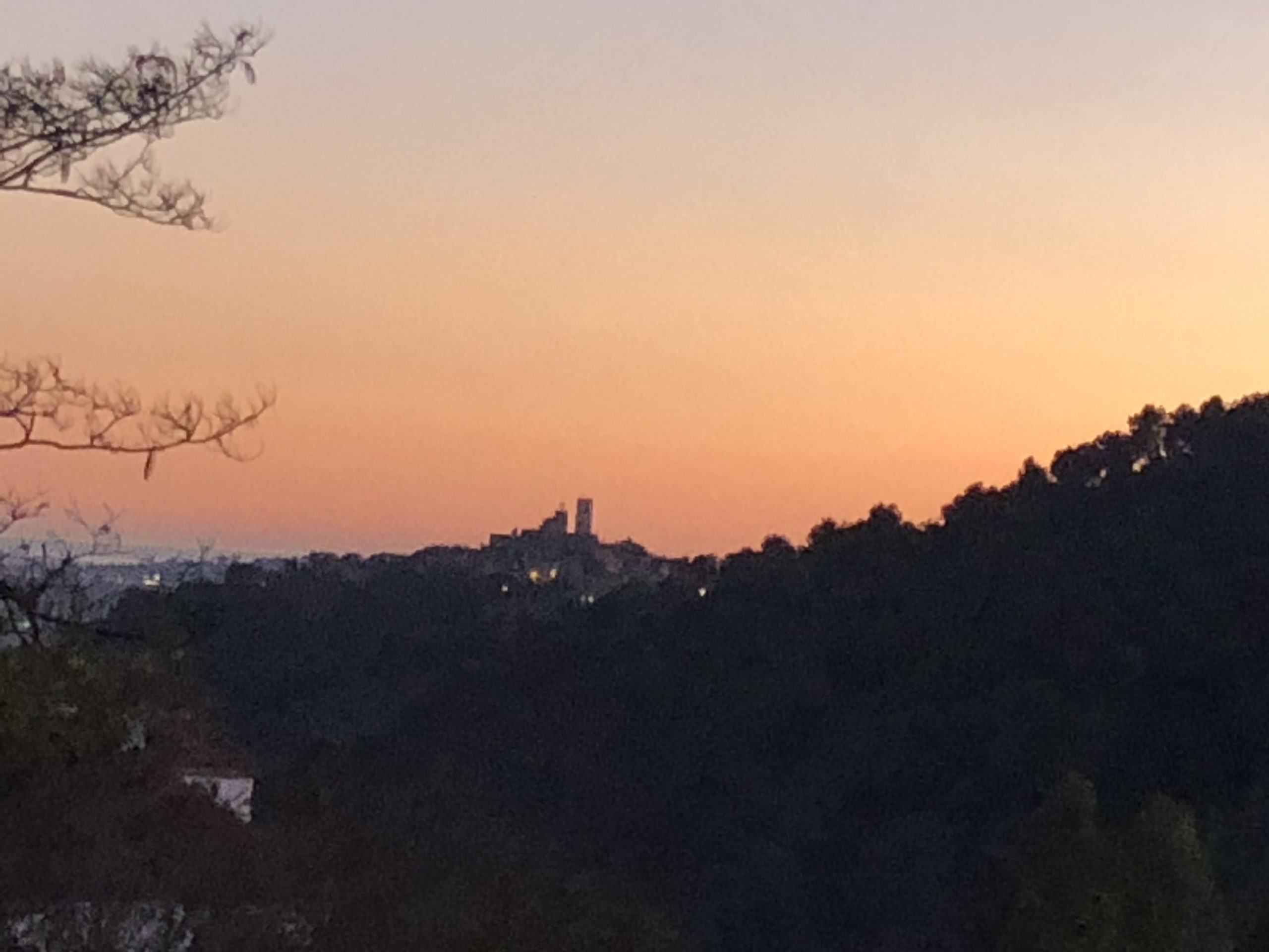 Solnedgang over en fjern by med silhuetter av åser og trær, og en pastellfarget himmel som går fra oransje til blå.